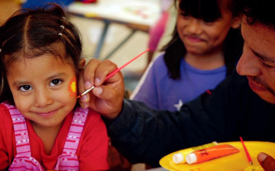 Parent applying facepaint to young child with older sibling looking on